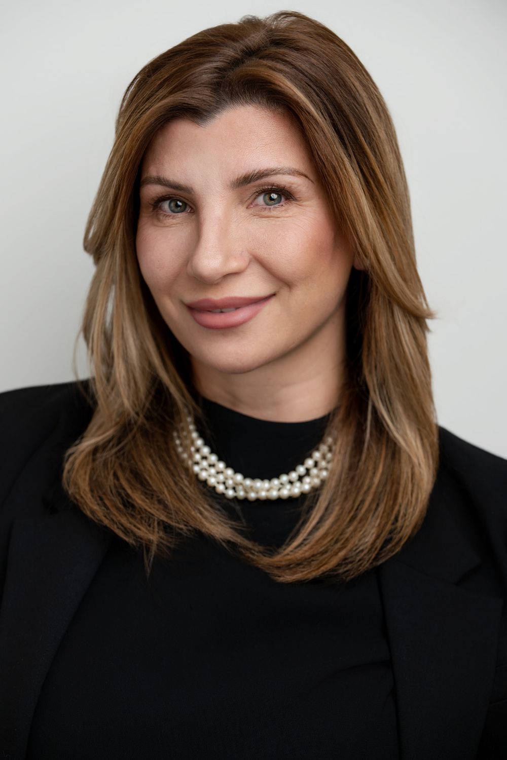 Professional corporate headshot portrait featuring brown hair, pearl necklace and black blazer against light background.