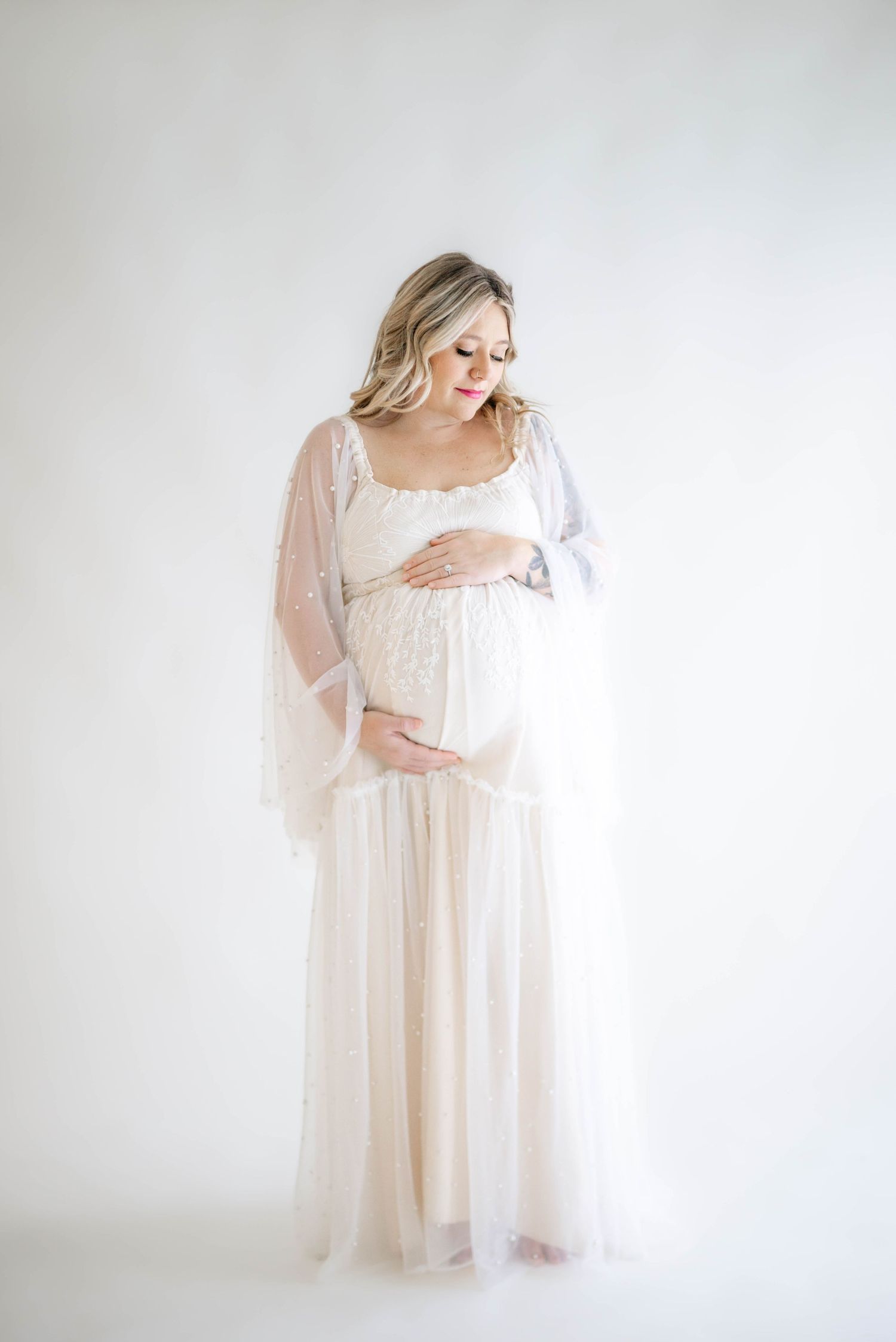 A person in a flowing white lace maternity gown cradles their baby bump against a light gray backdrop.