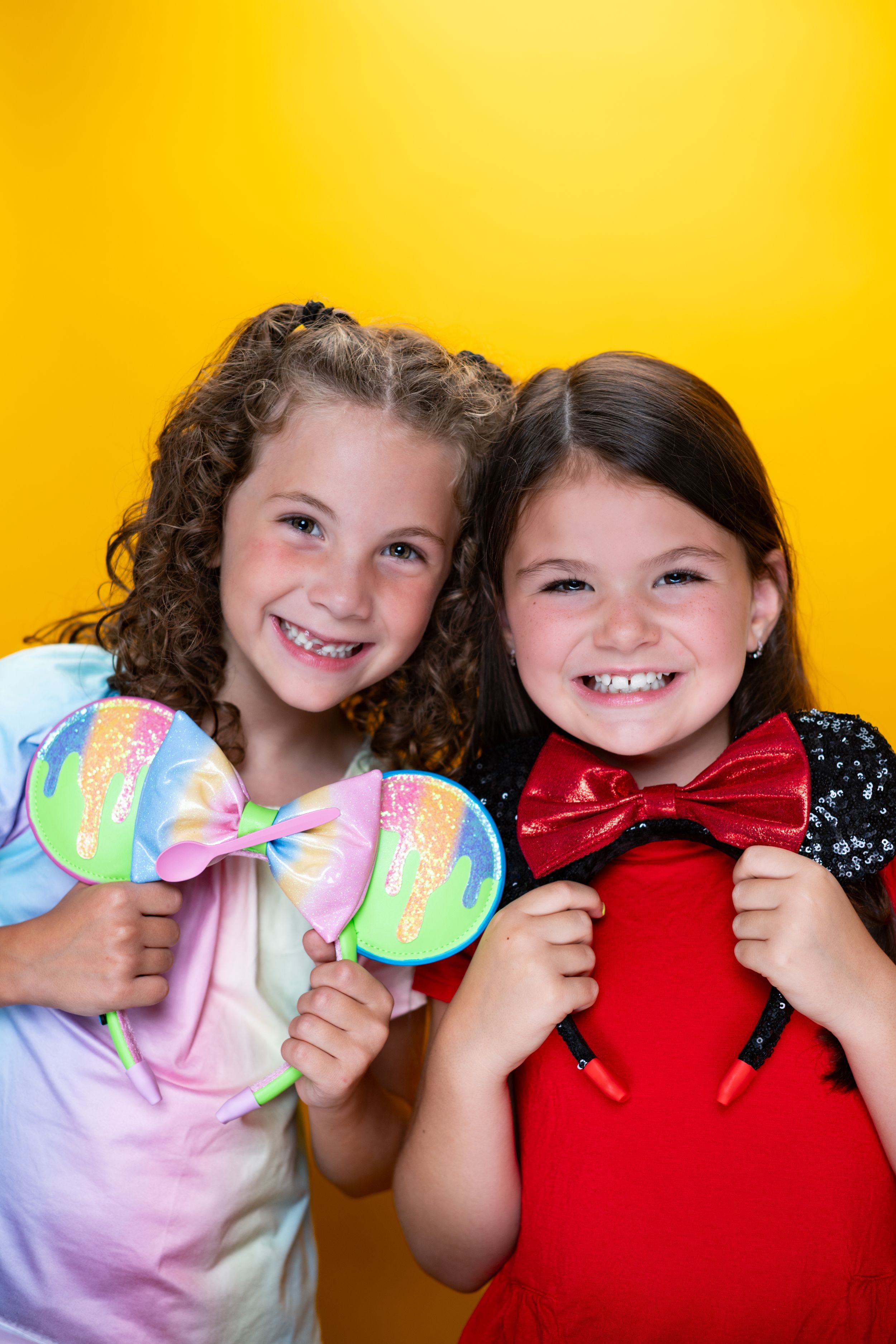 Two children wearing colorful bows holding Mickey Mouse ears against yellow background modeling for Hug A Band.