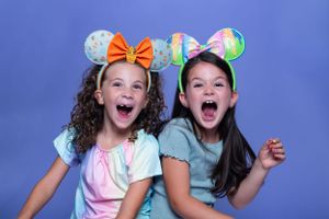 Two friends wearing Disney-themed ears striking playful poses against blue modeling for Hug A Band.
