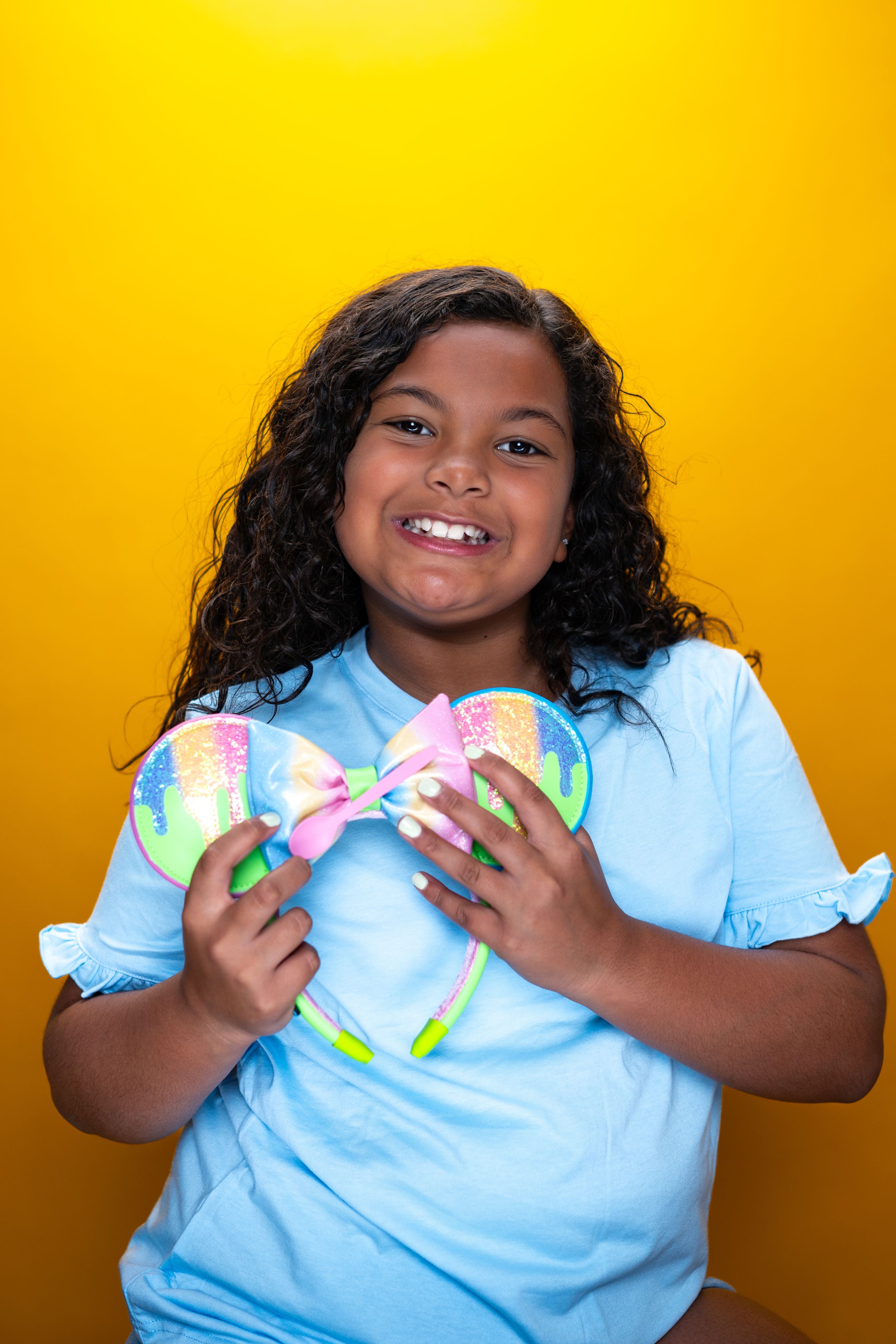 Child in blue shirt holding pastel bow headband against yellow background modeling for Hug A Band.