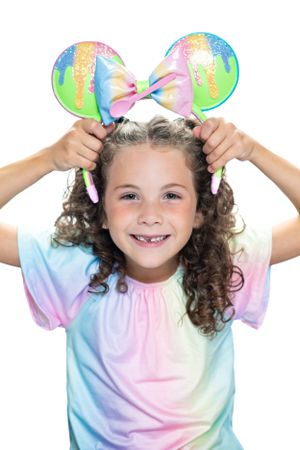 Child in tie-dye shirt wearing rainbow Mickey Mouse ears headband against white background modeling for Hug A Band.
