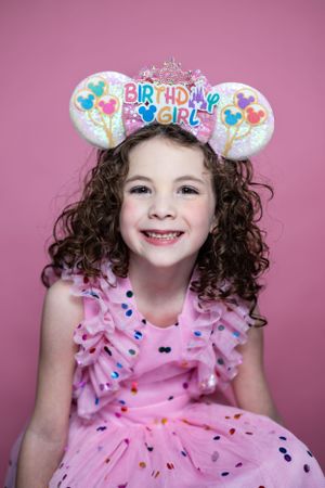 Child wearing pink birthday dress and Mickey Mouse ears with curly hair against pink background modeling for The Minnie Castle.