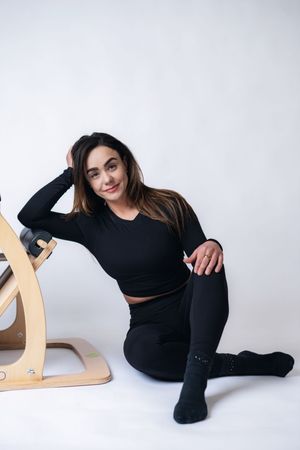 Woman in black activewear sitting on floor next to wooden Pilates chair, smiling in a white studio.