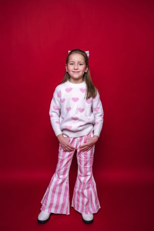 Young person in pink heart sweater and striped bell bottoms against red background wearing Sparkle In Pink.