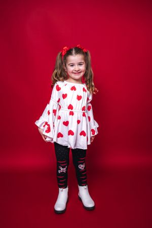 Child in heart print dress with red bows and white boots against red background.