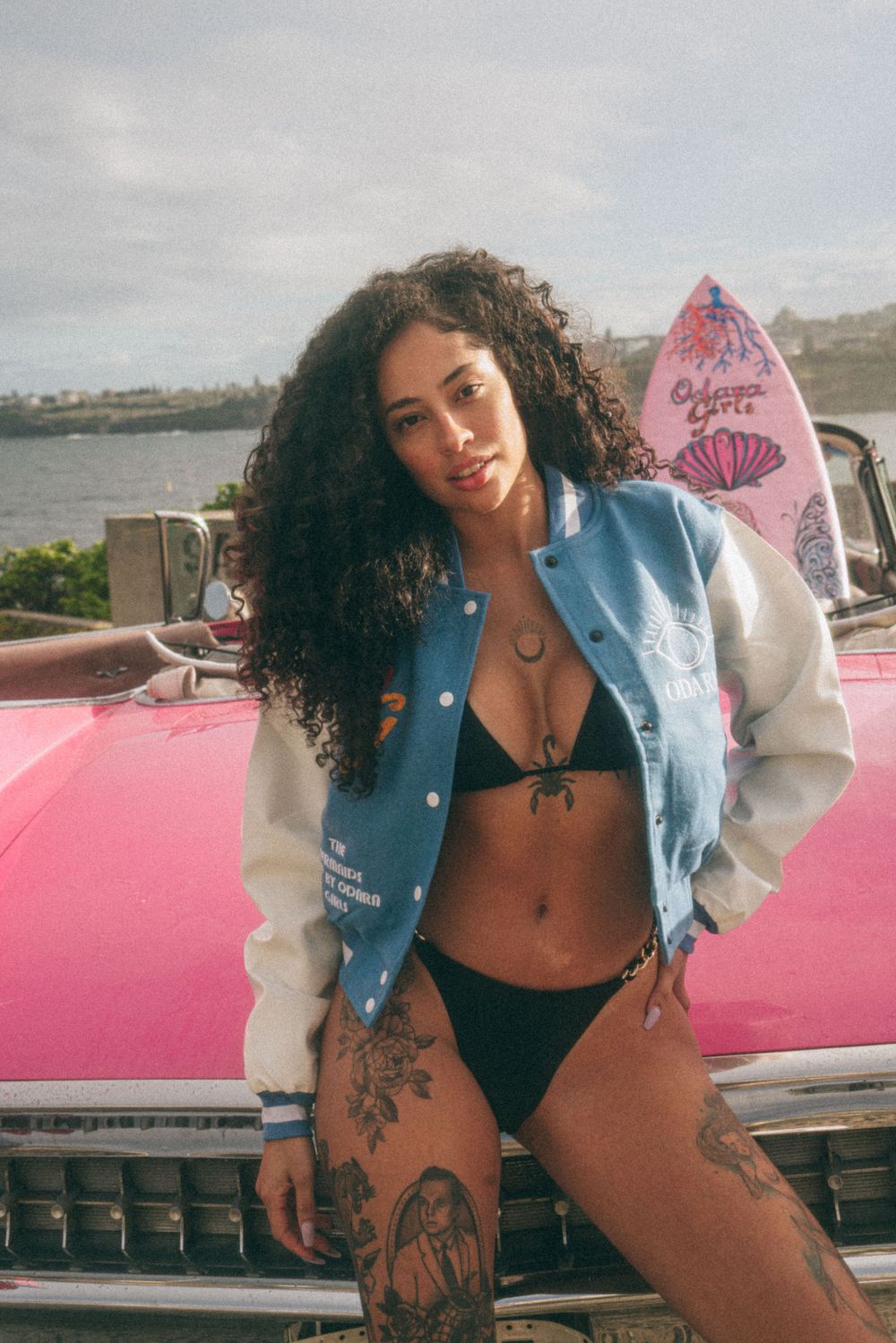 Model in varsity jacket poses with surfboard against pink classic car backdrop.