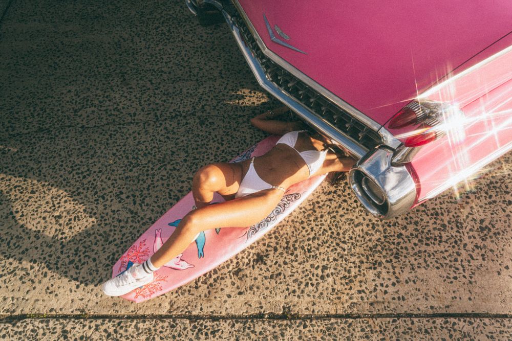 Model in swimwear lying on ground next to pink classic car.