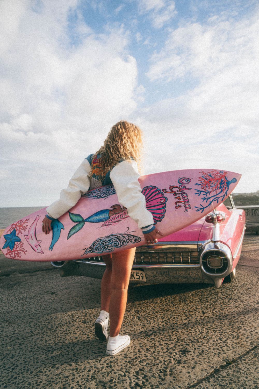 Person holding pink surfboard decorated with mermaids against vintage car.