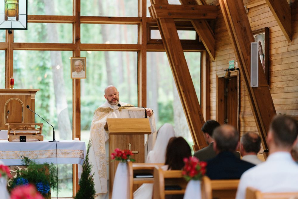 Interior of a rustic wooden chapel with large windows and wedding guests seated in pews.