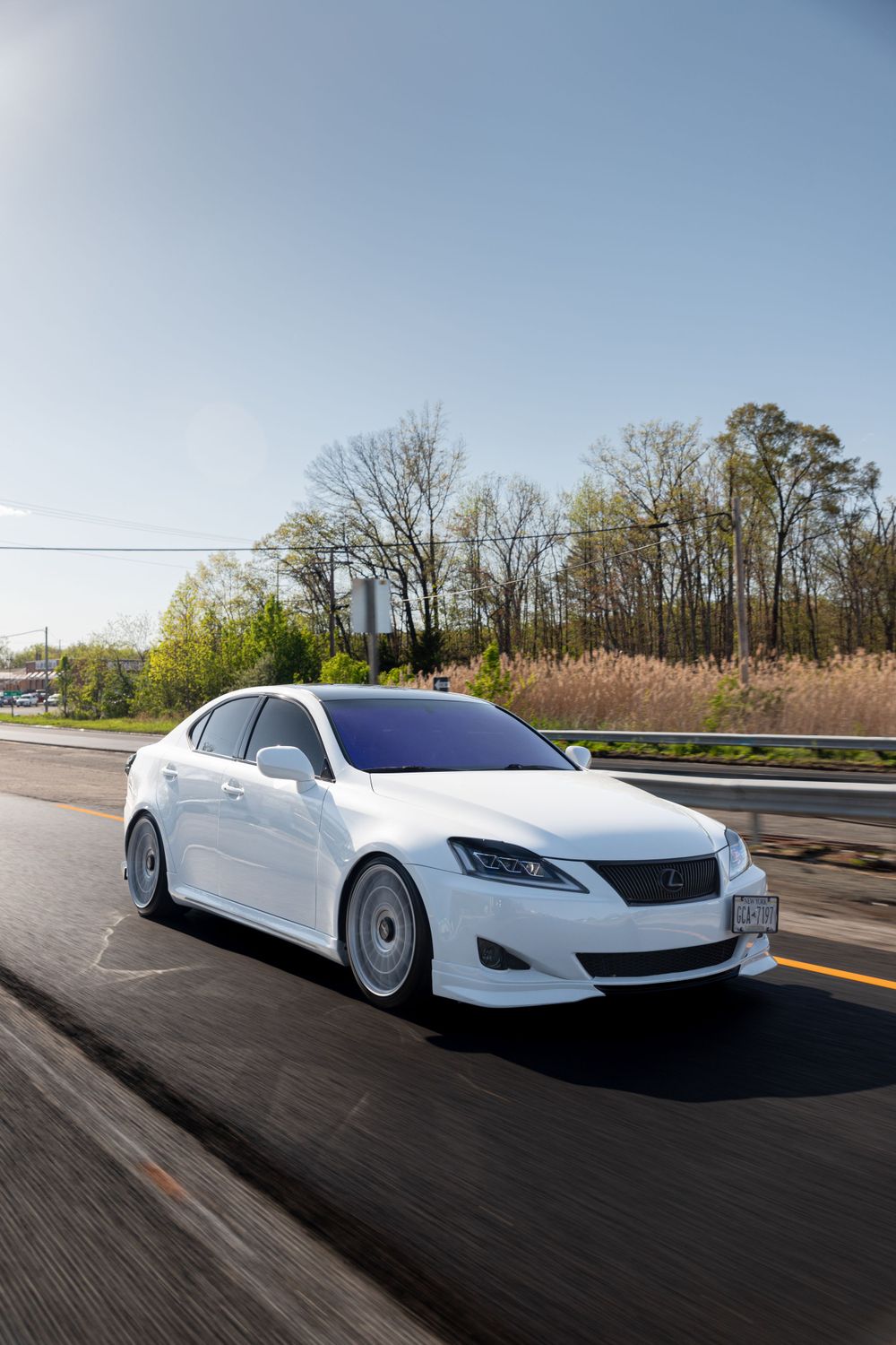 Lowered white Lexus sedan with custom wheels driving on a sunny road.