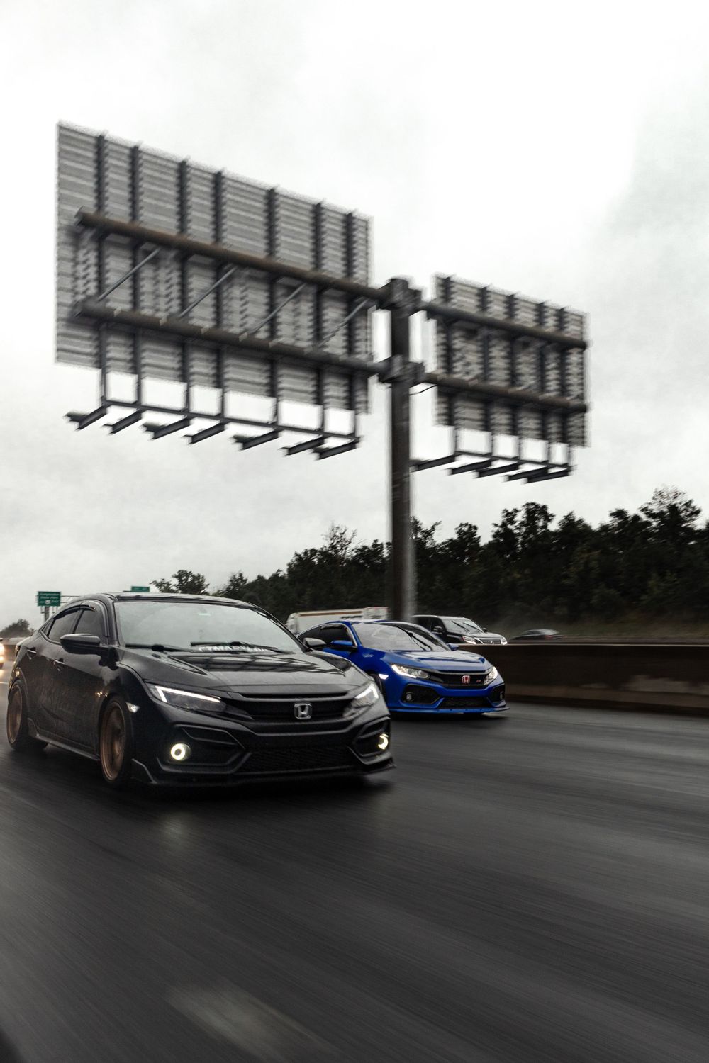 Two modified Honda Civics driving on highway beneath overhead signs.