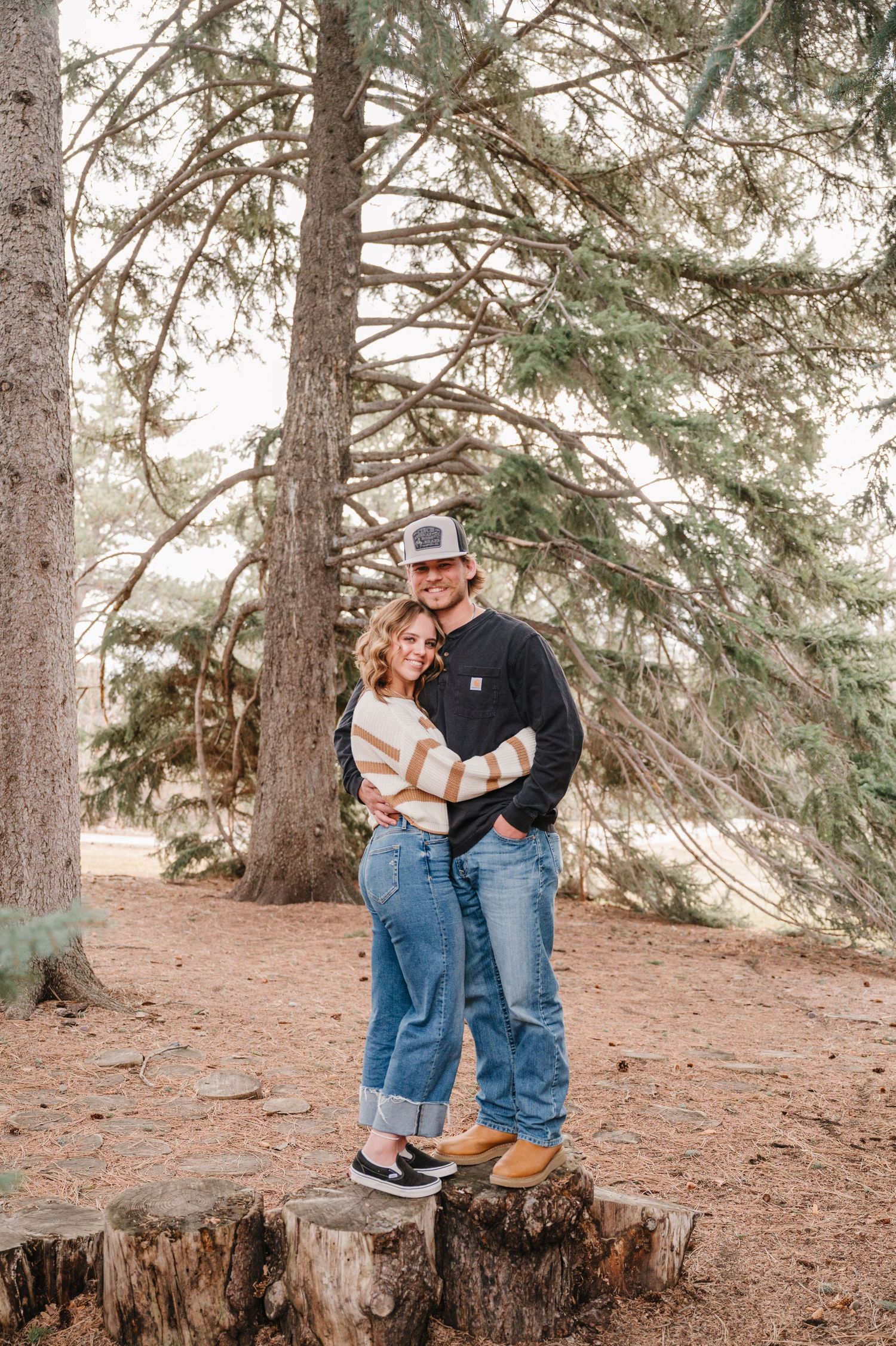 A couple stands together on a tree stump along a forest trail surrounded by tall evergreen trees.