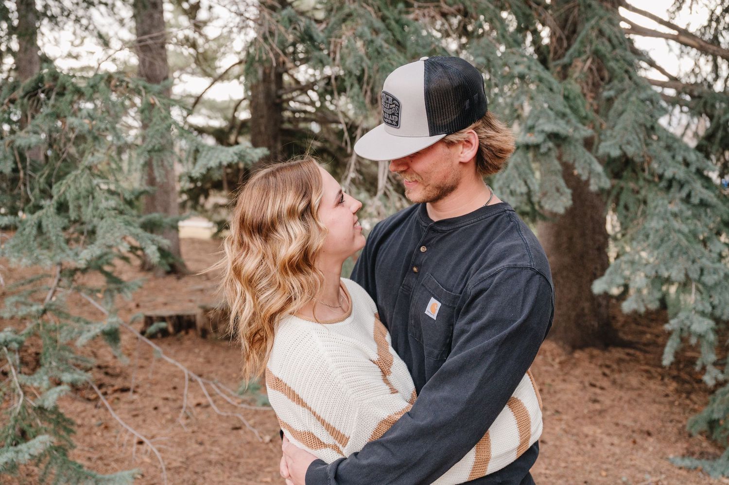 A romantic couple embraces outdoors among evergreen trees, wearing casual attire and looking lovingly at each other.
