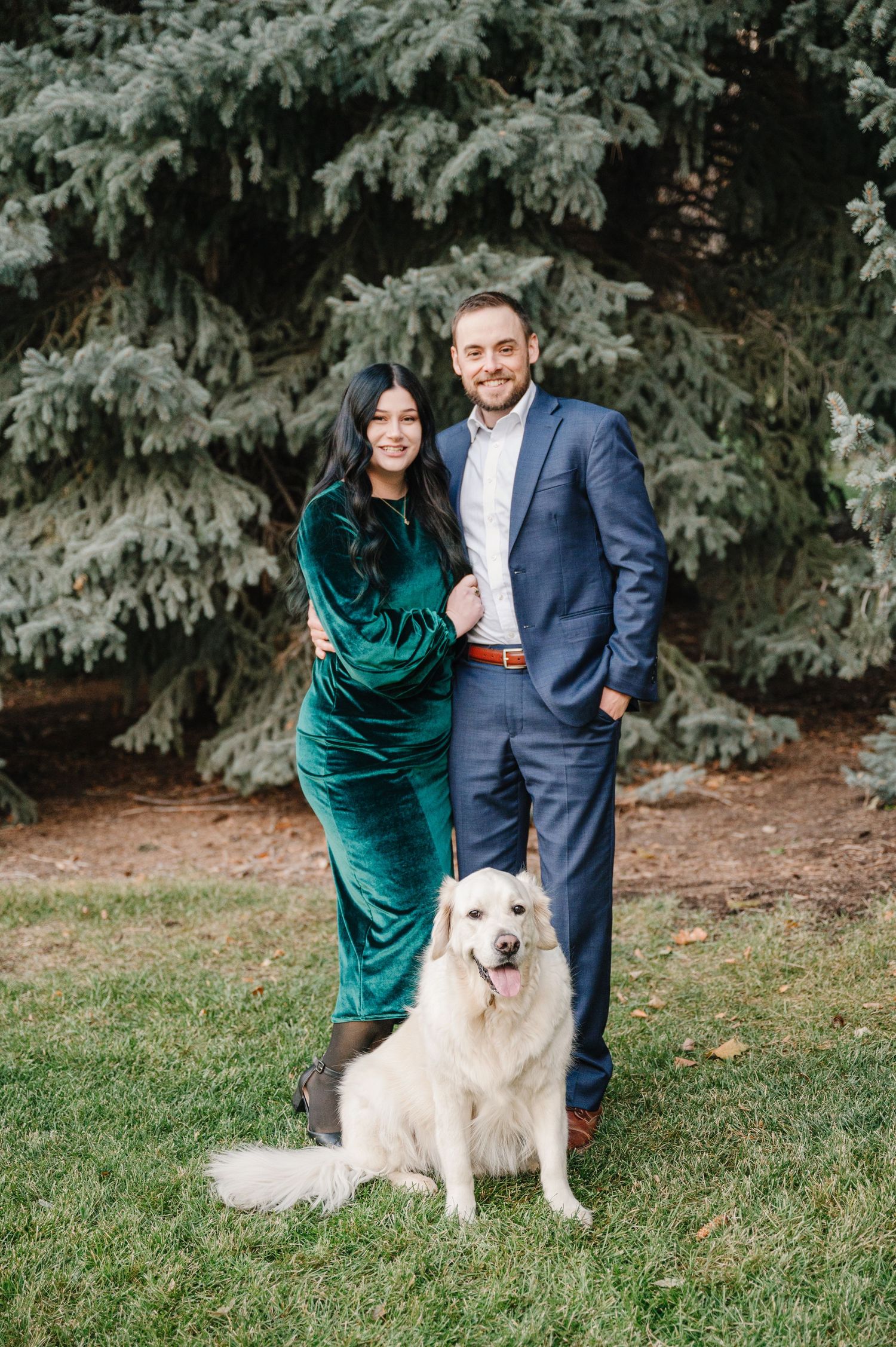 A couple in formal attire poses with their white dog against a snowy evergreen backdrop.