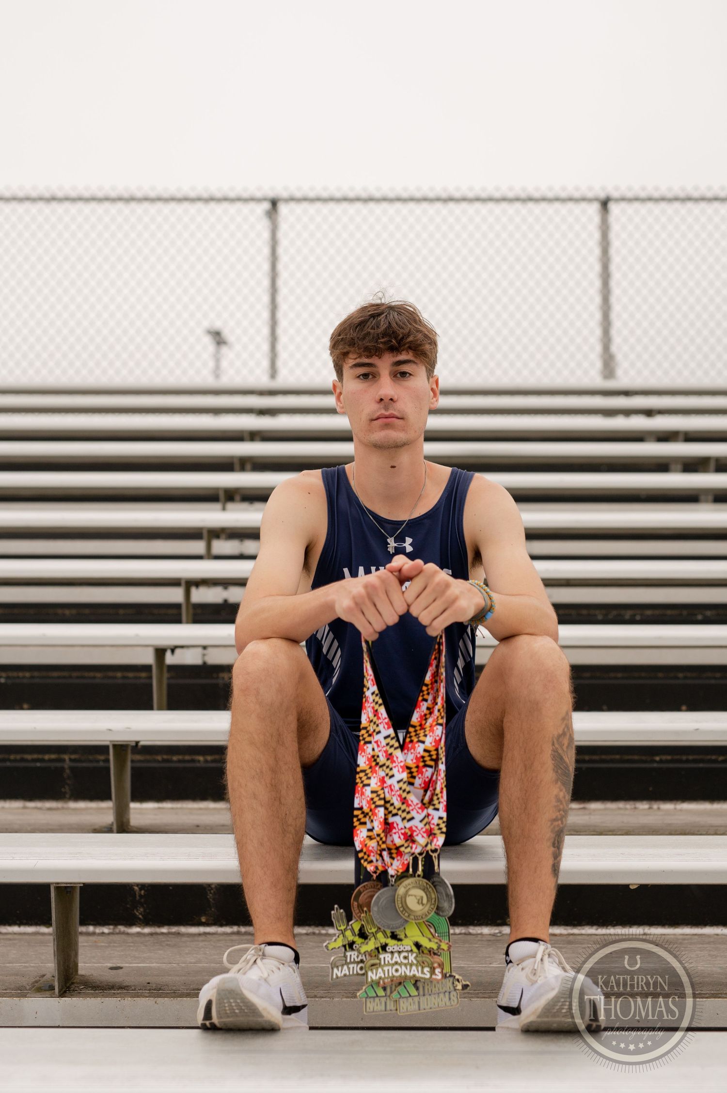 Track athlete poses with medals while sitting on metal bleachers under cloudy sky.