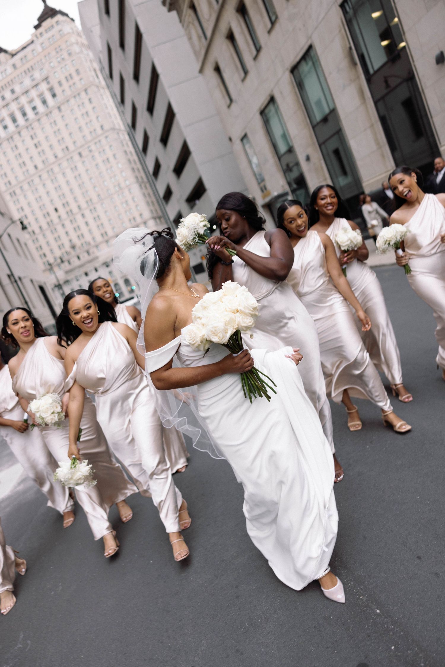 Bridal party in white dresses celebrates on city street with urban architecture backdrop.