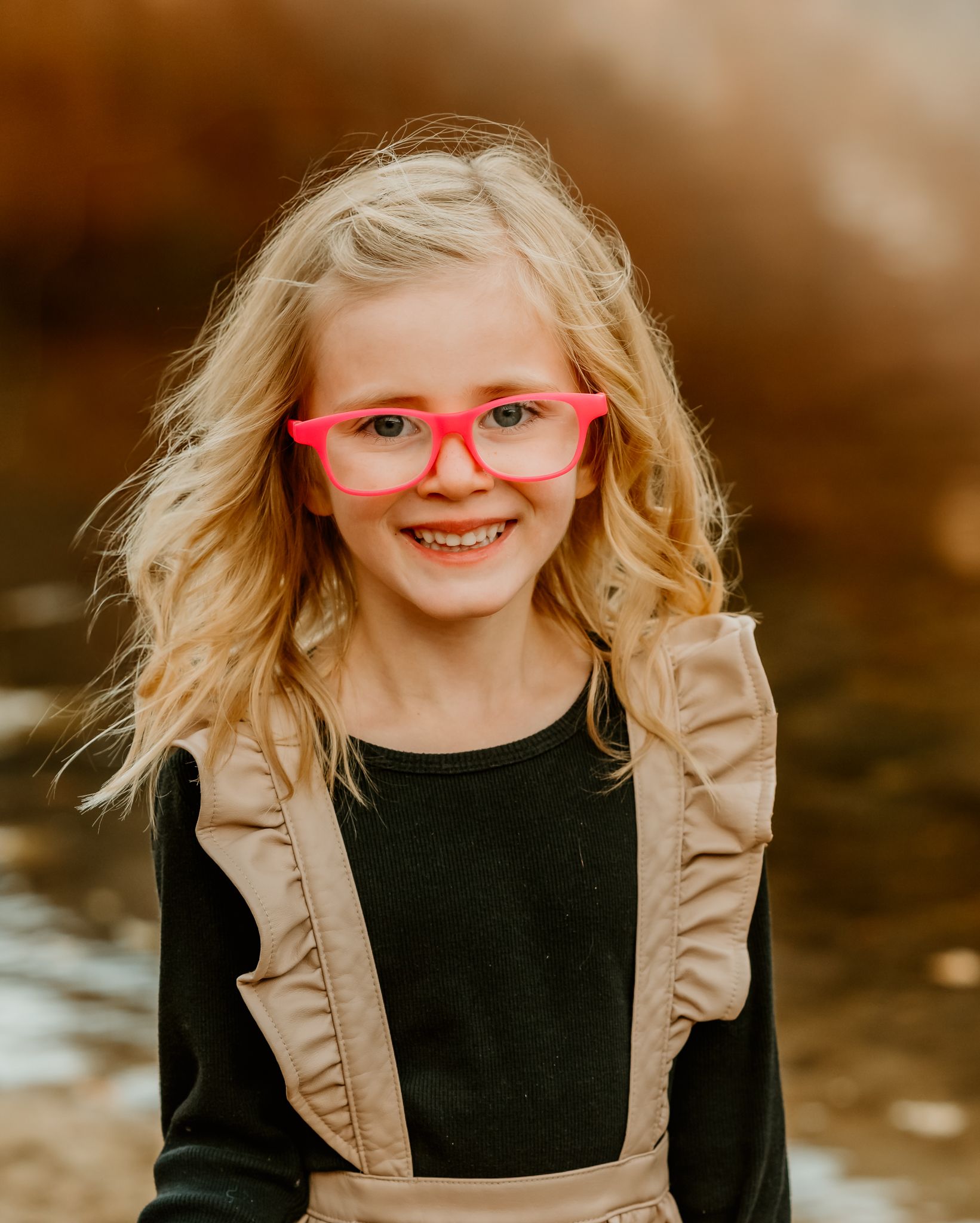 Child wearing bright red glasses and tan overalls smiles during outdoor portrait session.