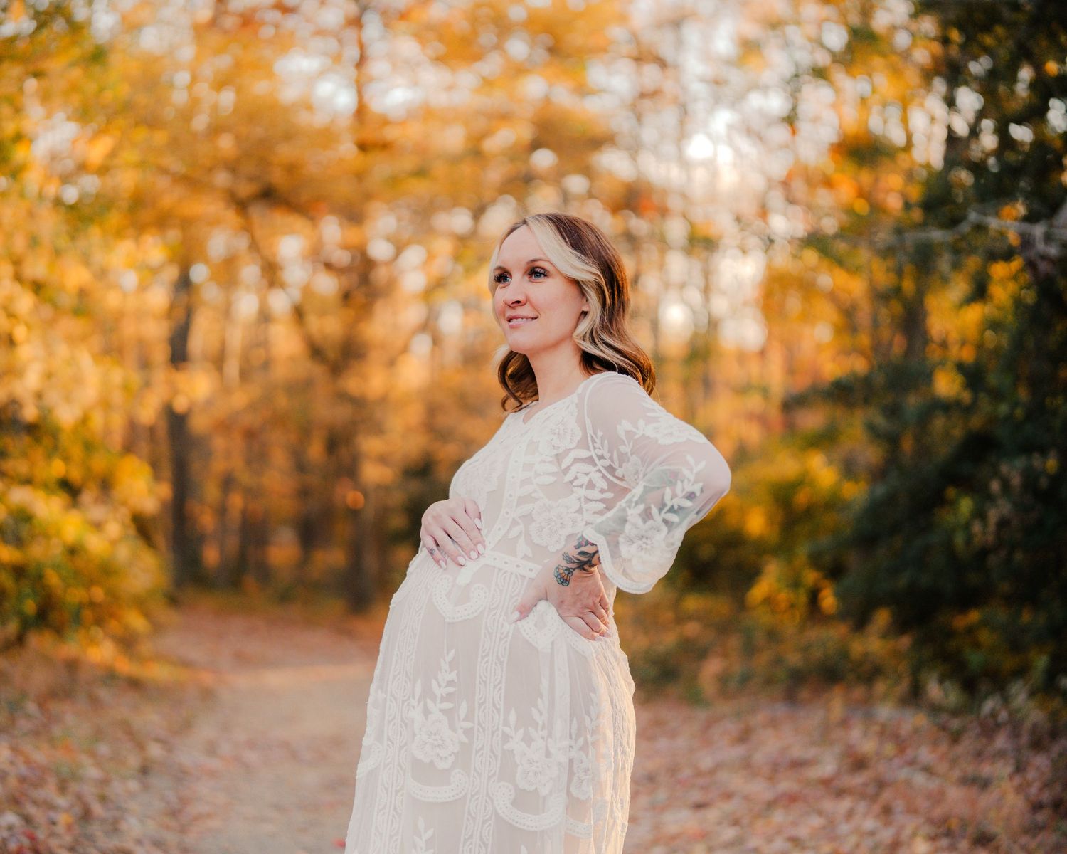 Person in white lace dress poses during autumn maternity photoshoot with golden foliage.