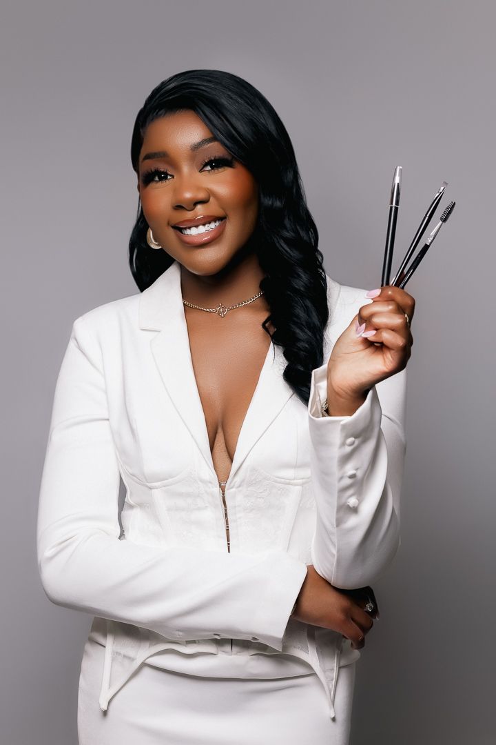 Professional makeup artist in white blazer holds makeup brushes while smiling against gray backdrop.