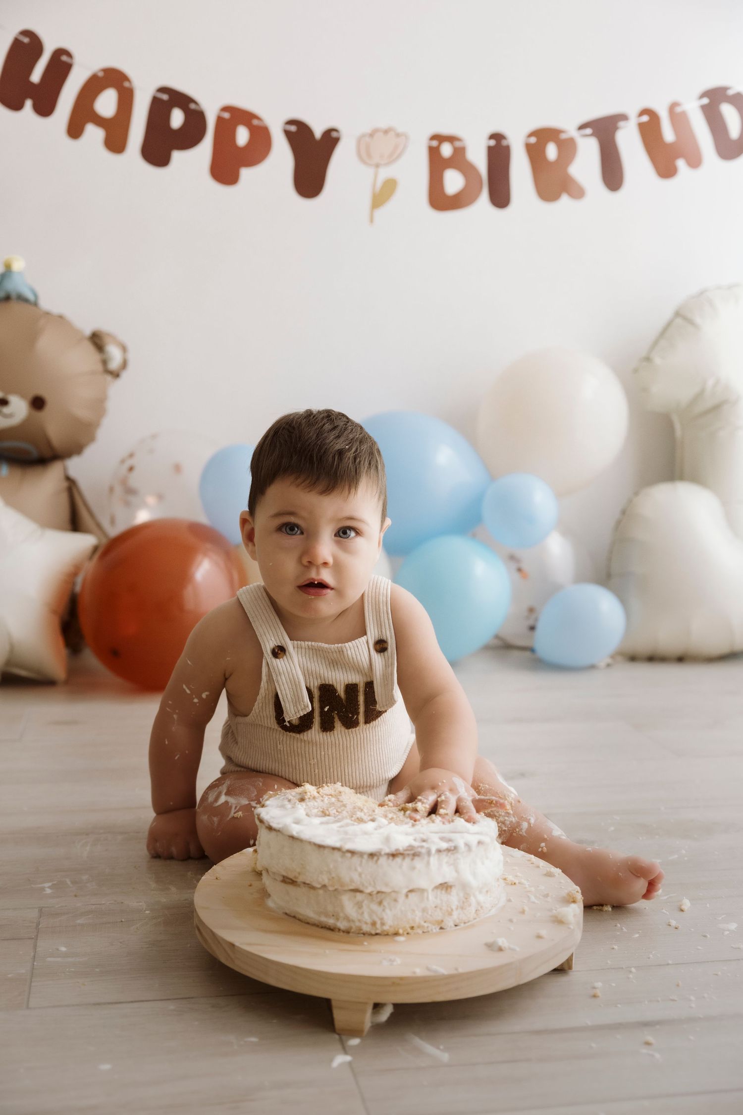 A cake smash photo session showing a baby in a black shirt exploring and playing with a white birthday cake.
