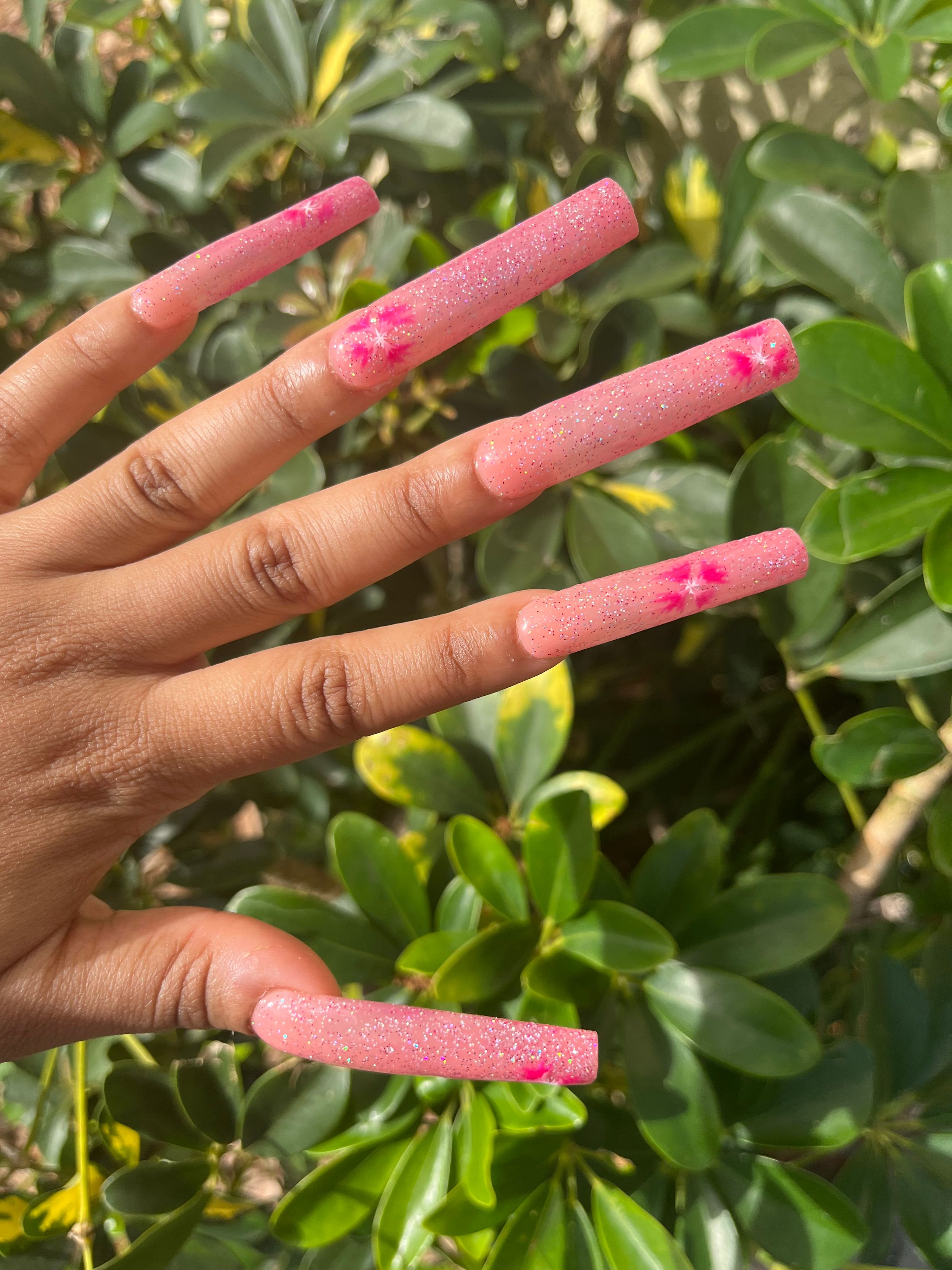 Long pink glitter acrylic nails displayed against green foliage background showing manicured hand with sparkly nail design.
