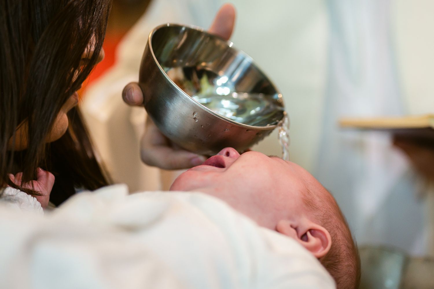 A baby lies on white fabric at a christening ceremony while holy water is poured from a silver vessel.