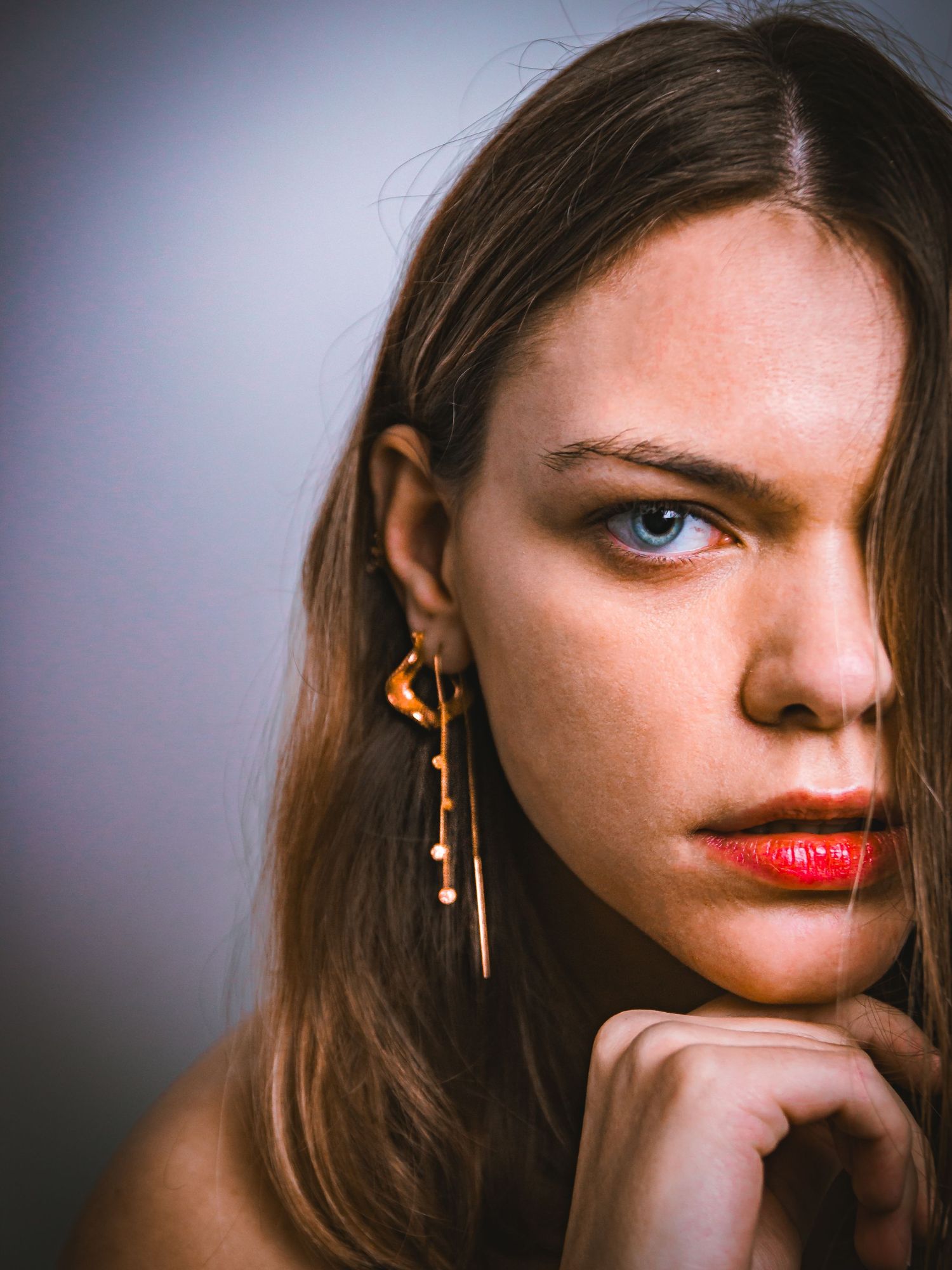 A model poses for a close-up portrait wearing distinctive gold dangle earrings and red lipstick against a moody backdrop.