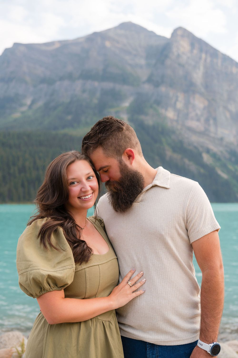 Couple embracing by turquoise mountain lake, woman in green dress smiling, bearded man in beige shirt, rocky peaks behind.