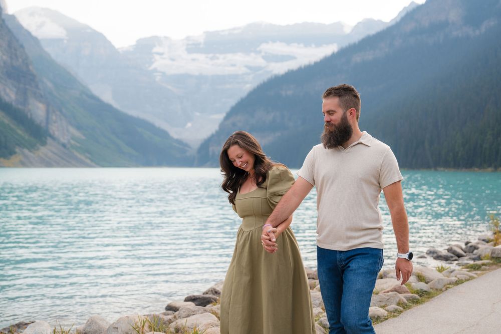 Couple walks hand-in-hand along turquoise Lake Louise shoreline with misty mountain glaciers in background.