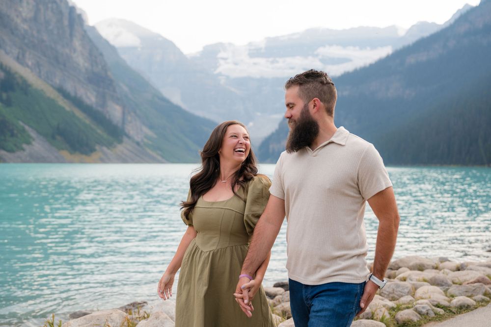 A laughing couple holds hands by turquoise Lake Louise with misty mountains in the background.