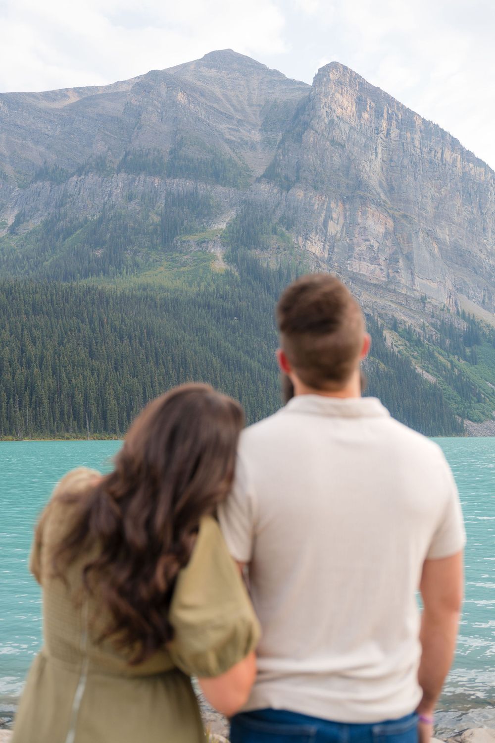 Couple standing together gazing at turquoise Lake Louise with dramatic Rocky Mountain peaks and forest backdrop.
