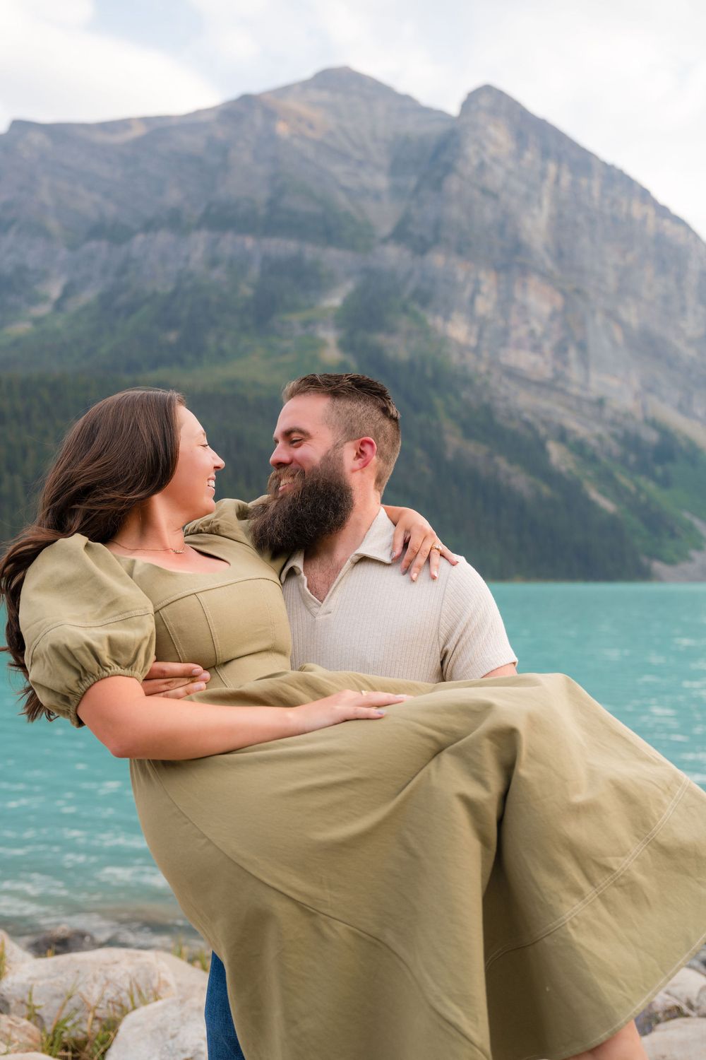 Man lifting woman in olive green dress at Lake Louise, both smiling with stunning Rocky Mountain scenery.