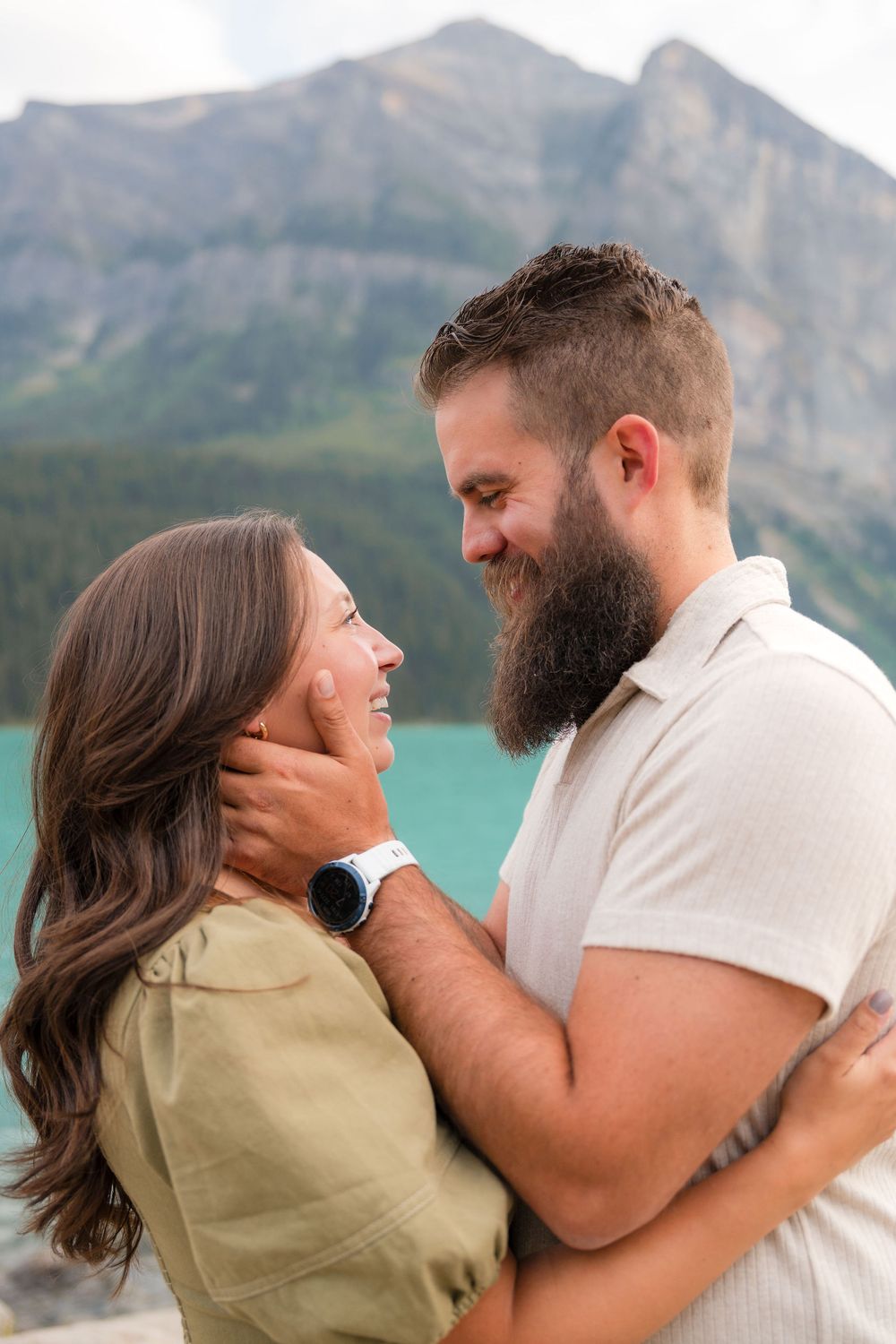Couple smiling and embracing near a turquoise mountain lake, man with beard holding woman's face tenderly.