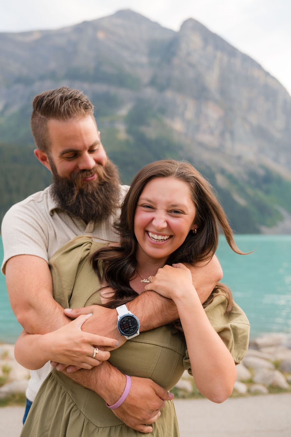 A bearded man hugs a laughing woman in a green dress near a turquoise lake with mountains in the background.