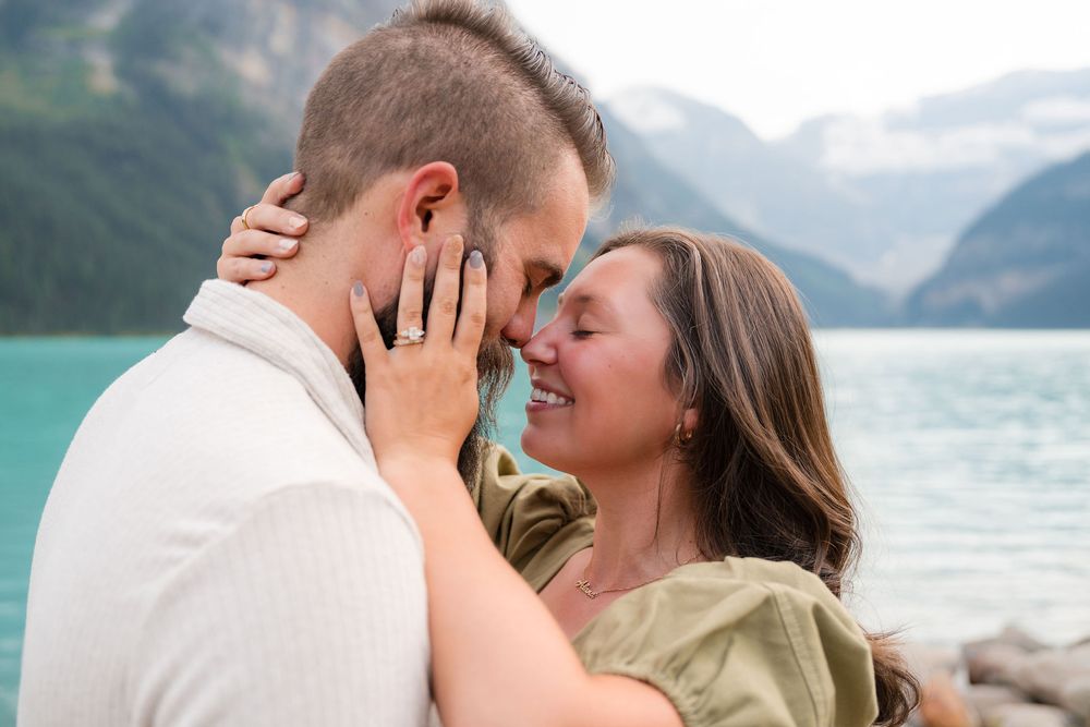 Couple sharing a romantic moment by turquoise Lake Louise with mountain backdrop, woman showing engagement ring.