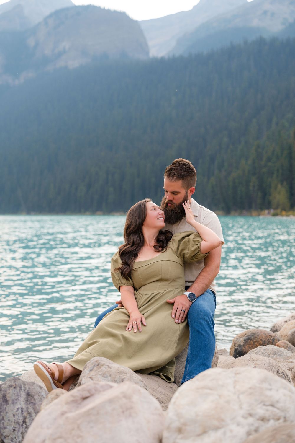 Couple sitting on rocks by a turquoise mountain lake, sharing a tender moment with forested peaks behind them.