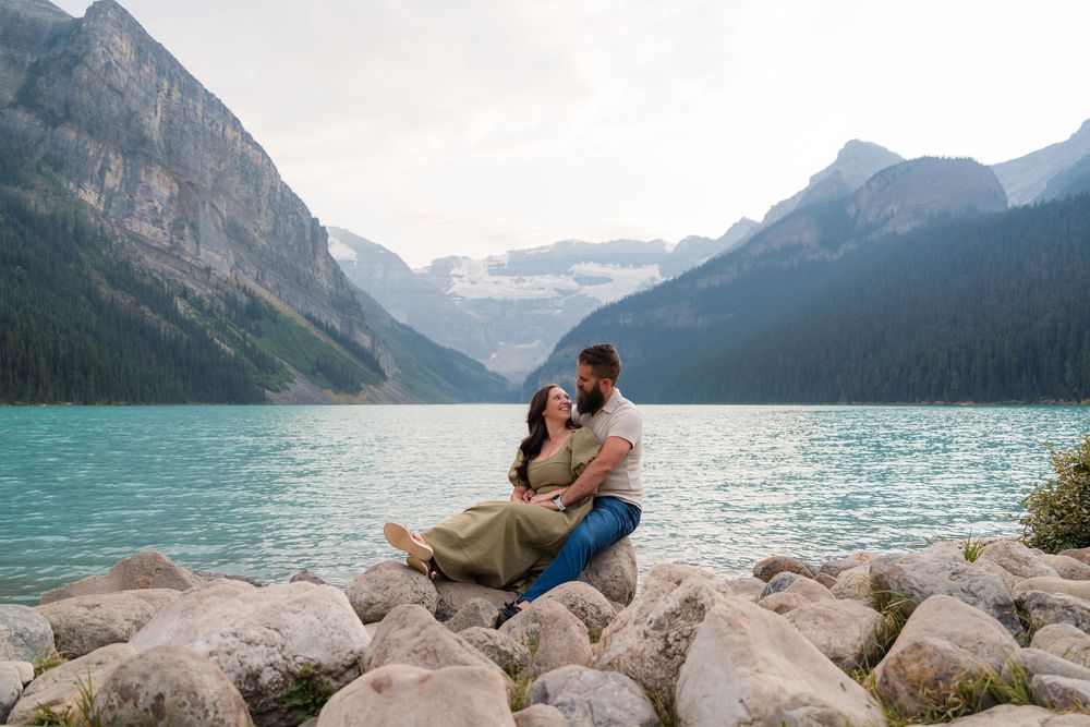 Couple sitting on rocks embracing by turquoise Lake Louise with stunning mountain backdrop in Banff National Park.