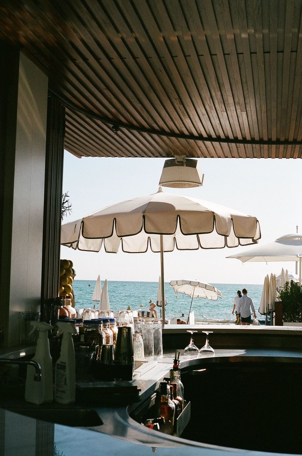 A beachside bar with tan umbrellas overlooking the ocean, featuring a wooden slatted ceiling and seaside panorama.