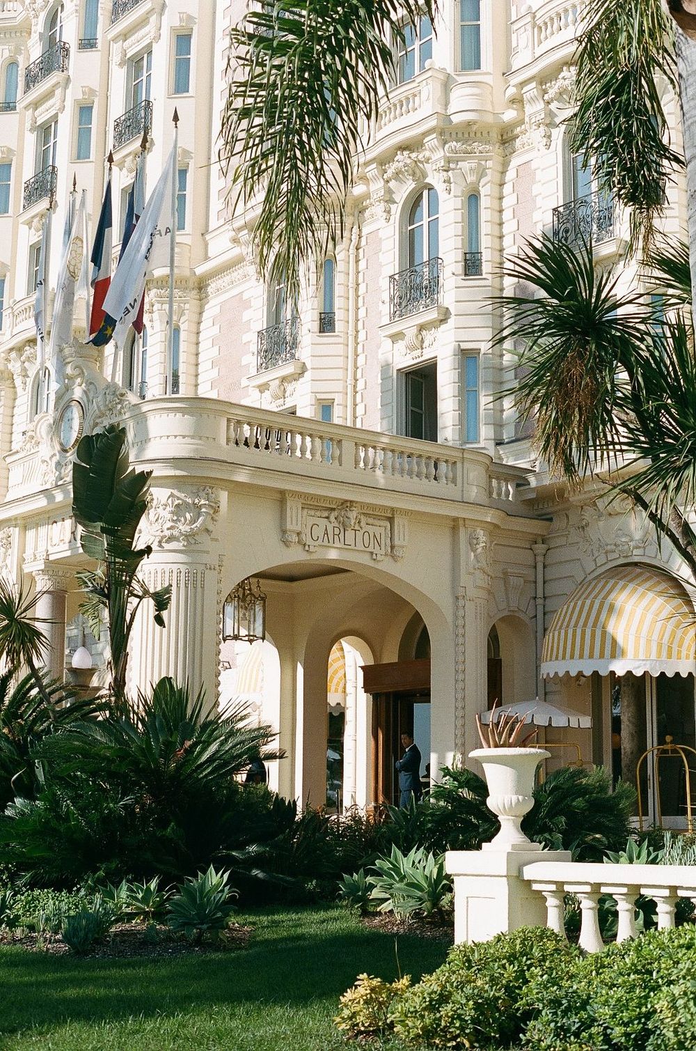 Elegant entrance of the Carlton Hotel in Cannes with ornate white architecture, palm trees and French flags.