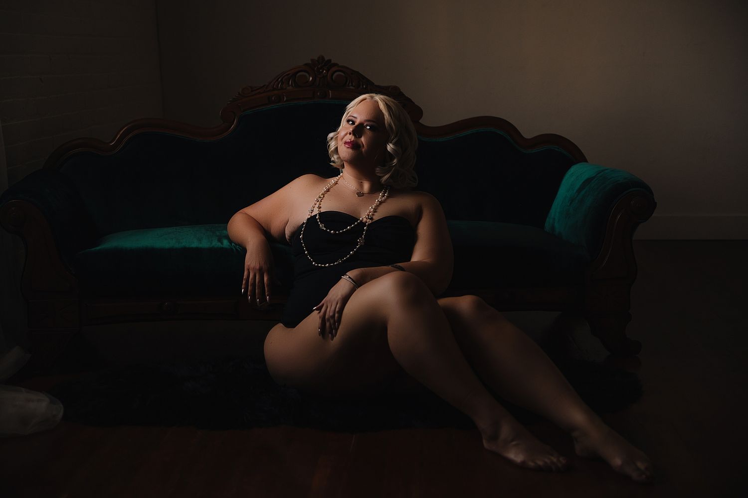 A dark and moody Utah boudoir photo of a woman  in a pearl necklace and black lingerie against a velvet teal couch.