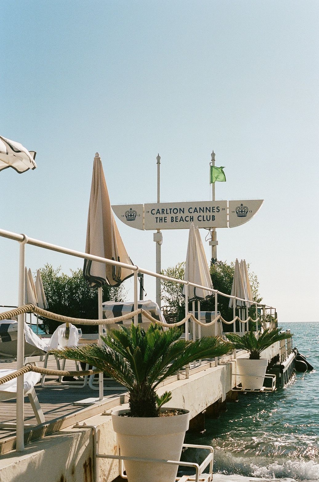 Luxury beachfront terrace at Carlton Cannes Beach Club with tan umbrellas and palm plants overlooking the Mediterranean Sea.
