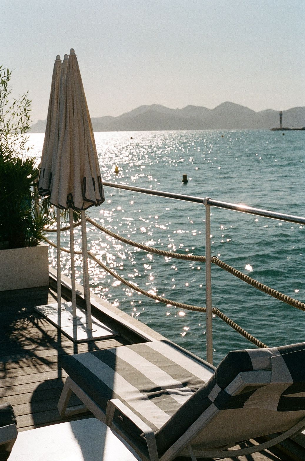 Ocean view deck with lounge chairs and folded umbrella overlooking sparkling Mediterranean waters and distant mountains.