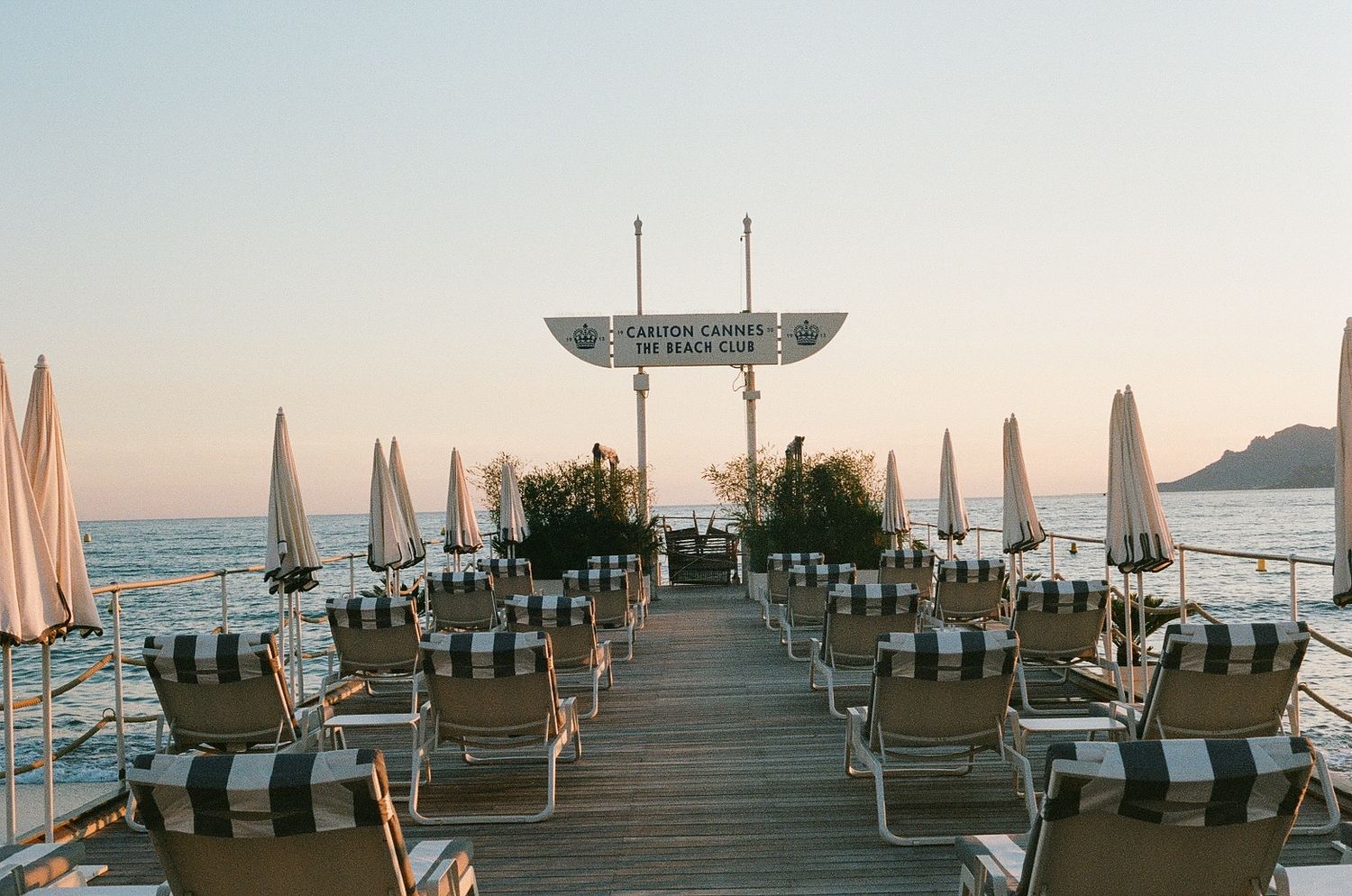 Beachfront wooden pier with striped lounge chairs and closed umbrellas at sunset overlooking the Mediterranean Sea.