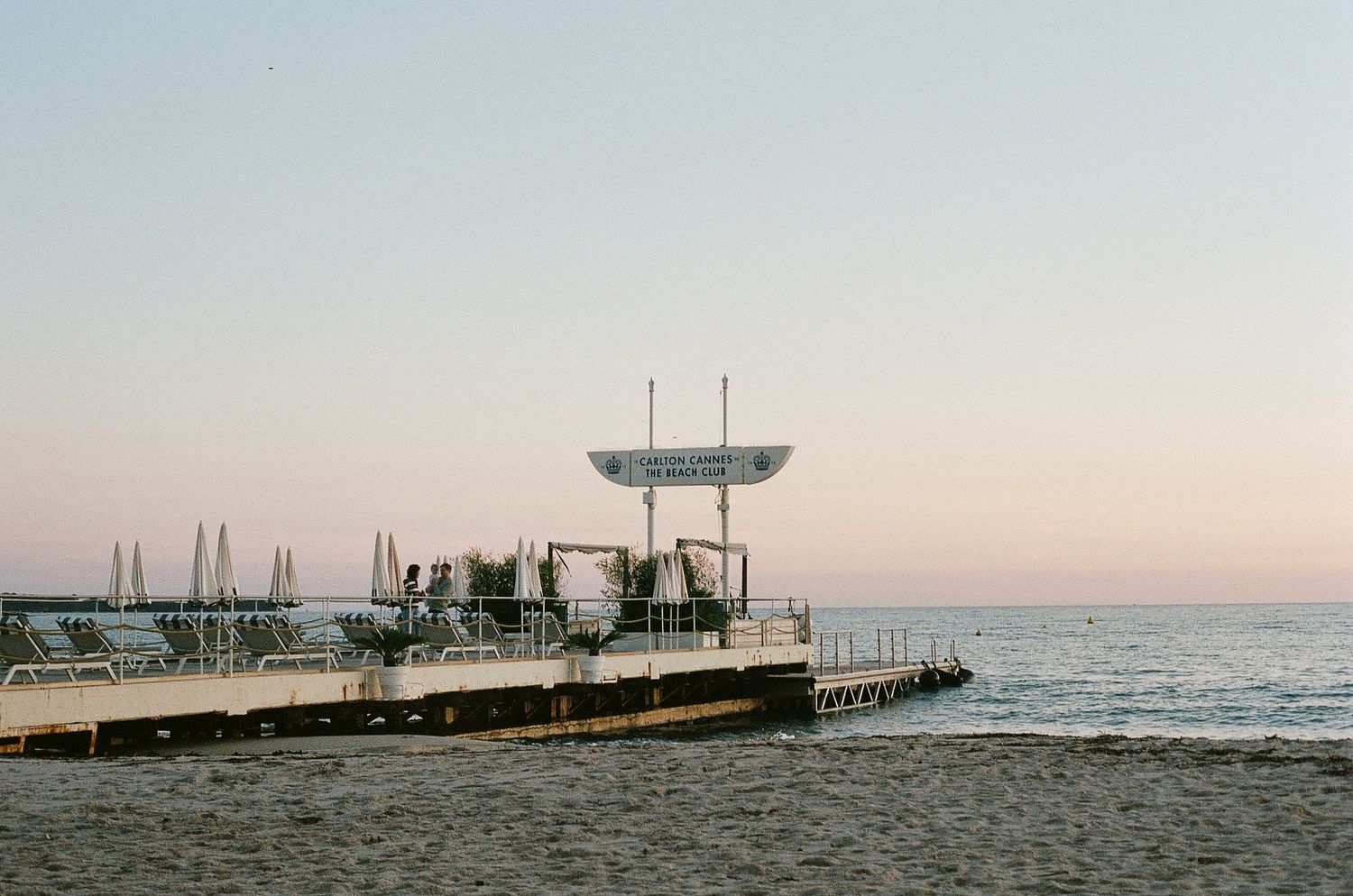 A lifeguard tower stands on a serene beach pier during sunset with silhouetted boats in the distance.