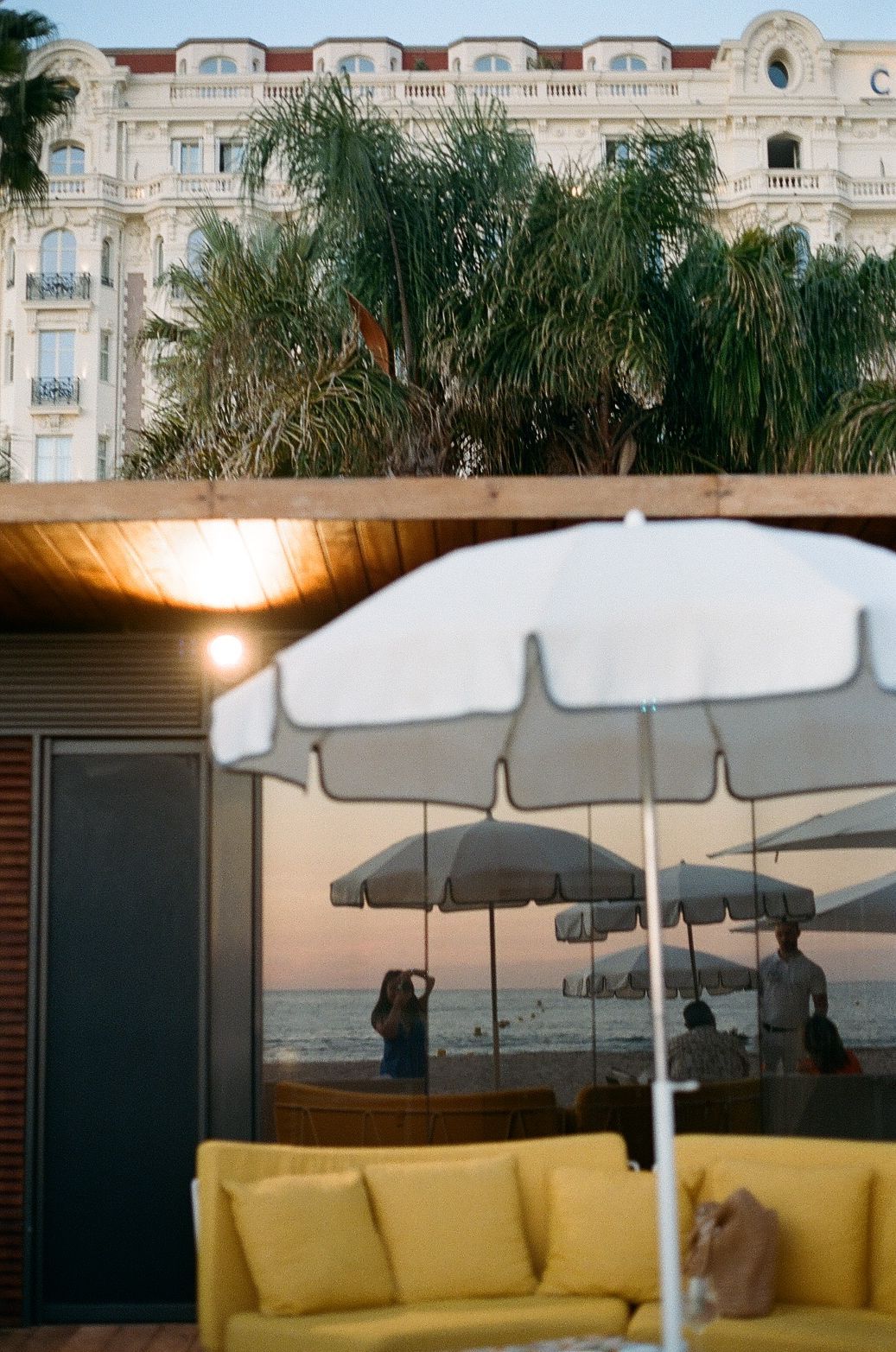 Luxury hotel terrace with yellow couch and white umbrellas overlooking the sea at sunset, with ornate facade in background.