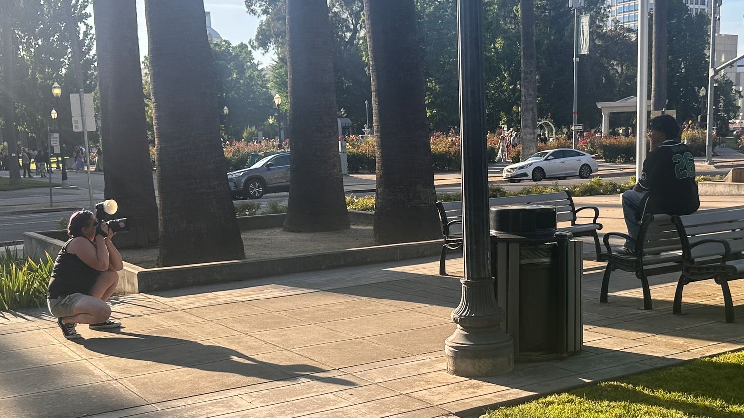 Person crouching to photograph urban park scene with trees, benches, and trash can on sunny day.