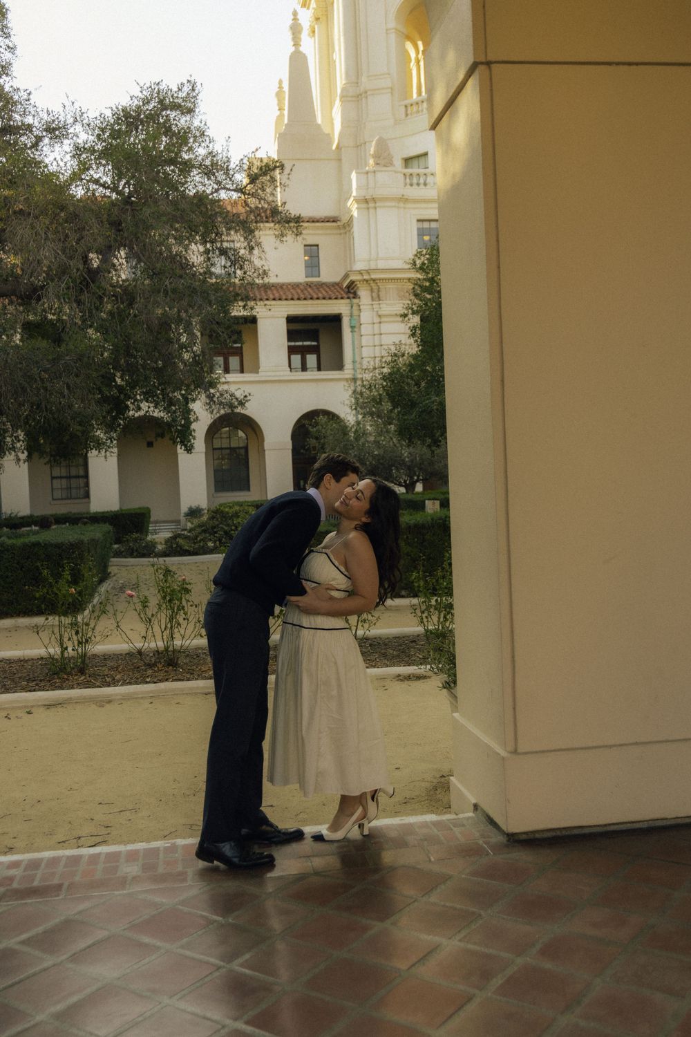 A couple shares an intimate moment near a cream-colored building with arched windows.