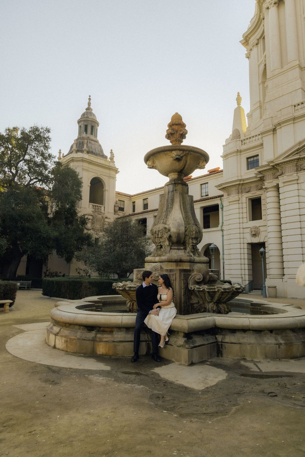 A couple sits by an ornate fountain in front of a historic building with Spanish colonial architecture.