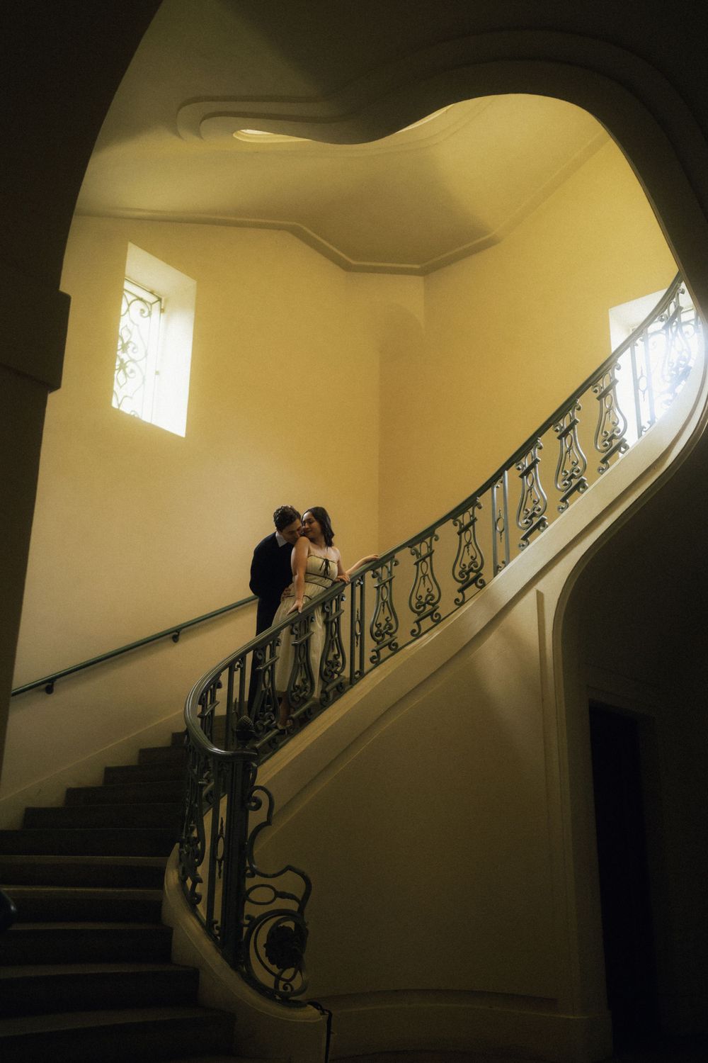 A silhouetted couple stands on an elegant curved staircase with ornate railings in a dimly lit historic building.